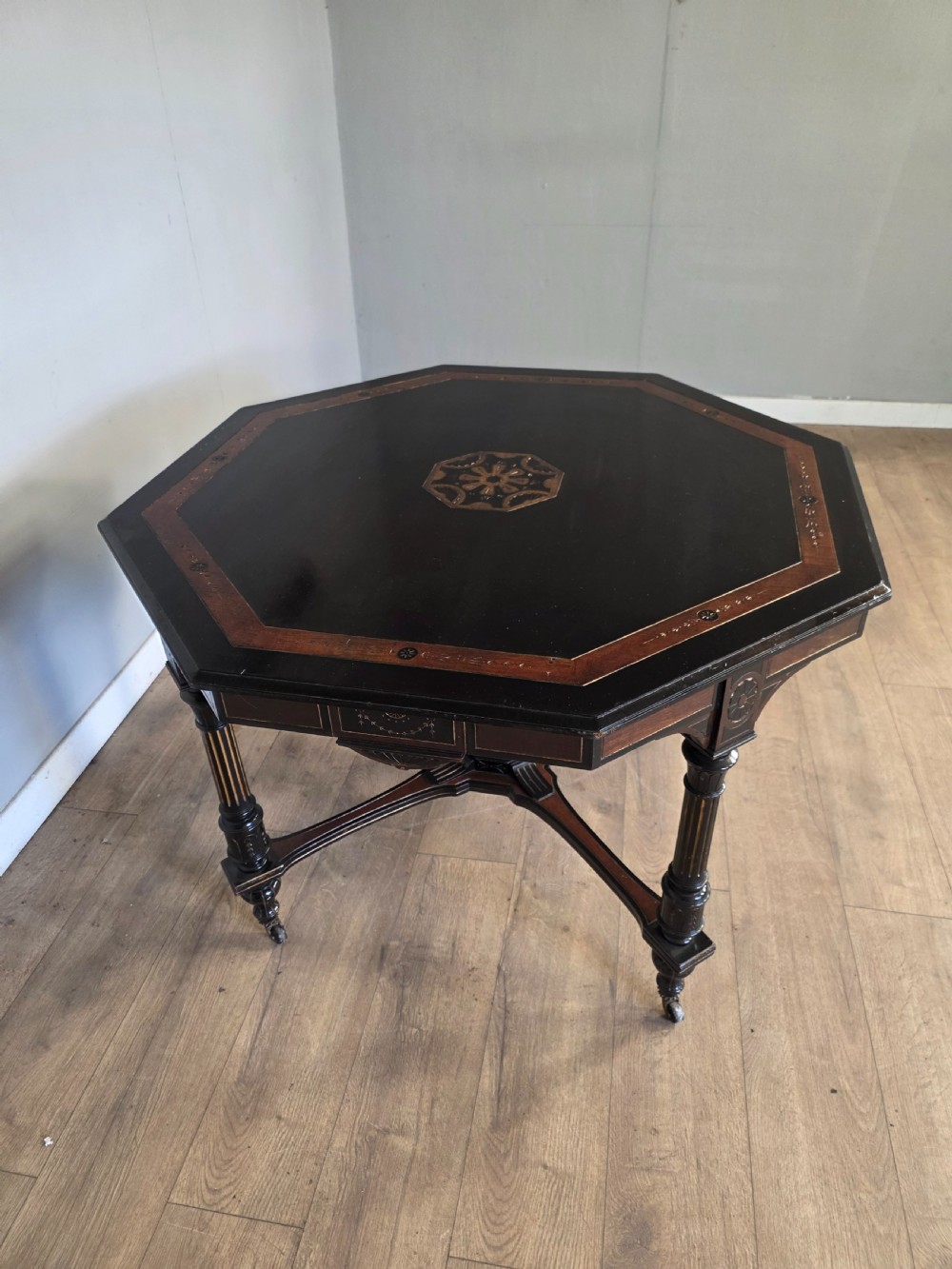 An antique Victorian octagonal centre table featuring an ebonised black finish, gilded accents, turned legs, an X-frame stretcher with a central finial, and brass castors. View VI