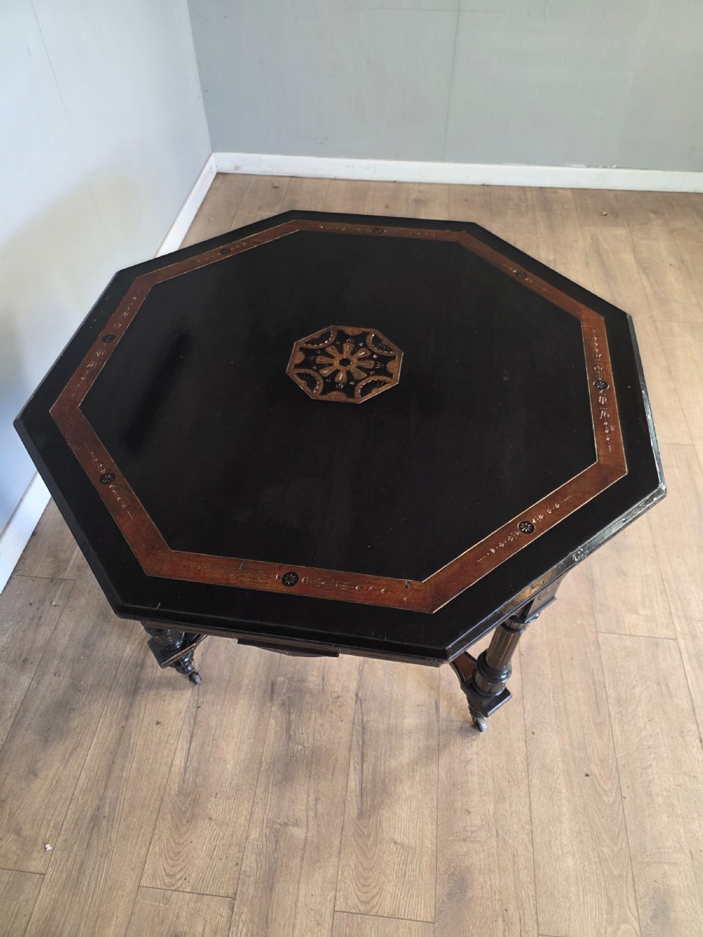 An antique Victorian octagonal centre table featuring an ebonised black finish, gilded accents, turned legs, an X-frame stretcher with a central finial, and brass castors. View VII