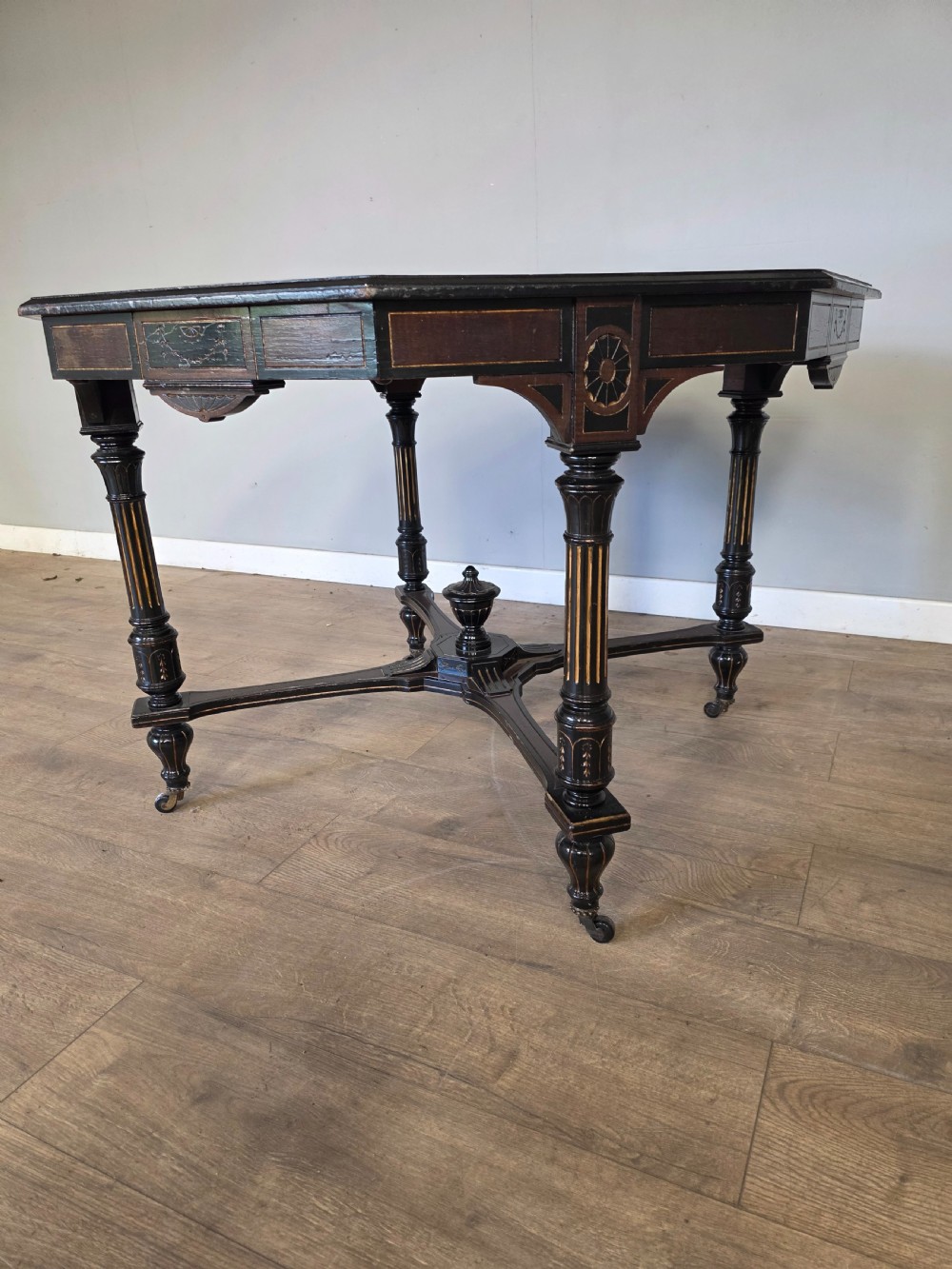 An antique Victorian octagonal centre table featuring an ebonised black finish, gilded accents, turned legs, an X-frame stretcher with a central finial, and brass castors. View IX