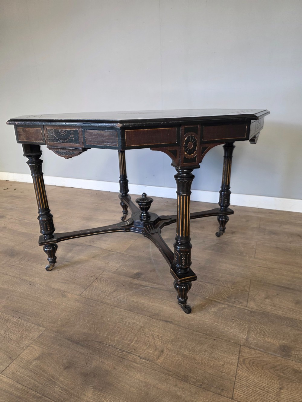 An antique Victorian octagonal centre table featuring an ebonised black finish, gilded accents, turned legs, an X-frame stretcher with a central finial, and brass castors. View X