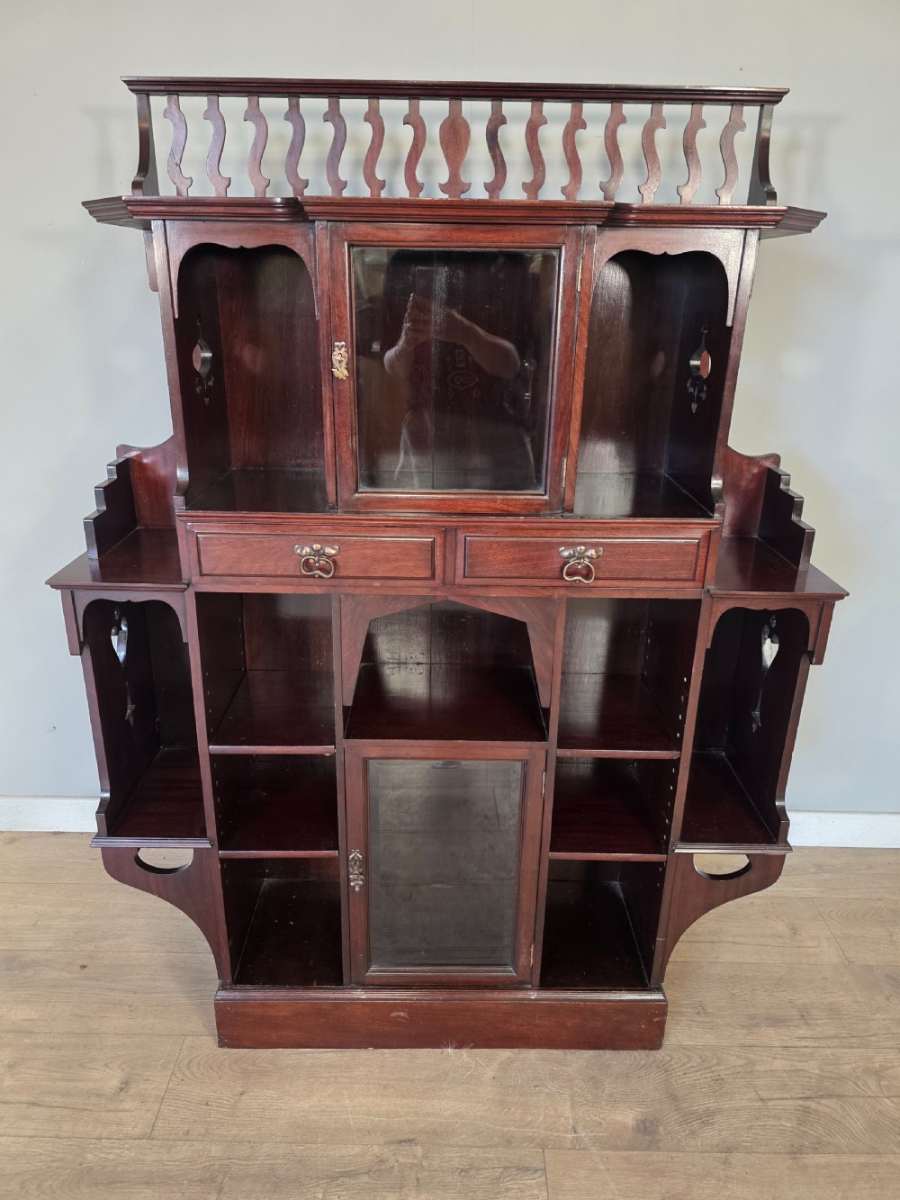 An antique dark mahogany Arts and Crafts display bookcase featuring a top galleried balustrade, open side shelves with pierced cut-outs, two glazed cabinet doors, and two middle drawers. View II
