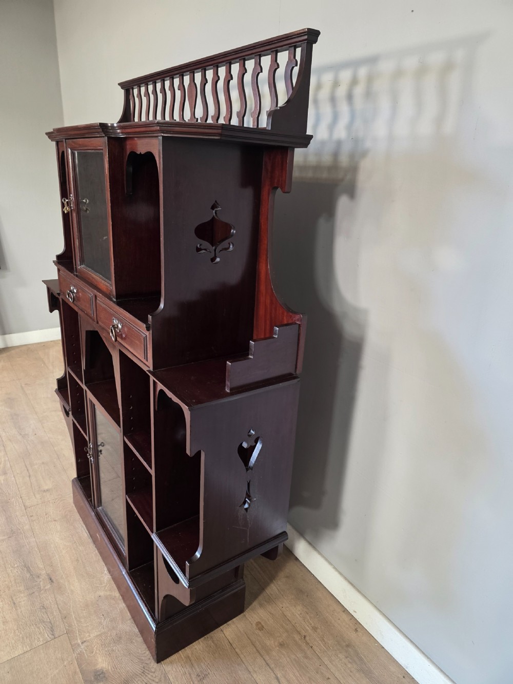 An antique dark mahogany Arts and Crafts display bookcase featuring a top galleried balustrade, open side shelves with pierced cut-outs, two glazed cabinet doors, and two middle drawers. View VI