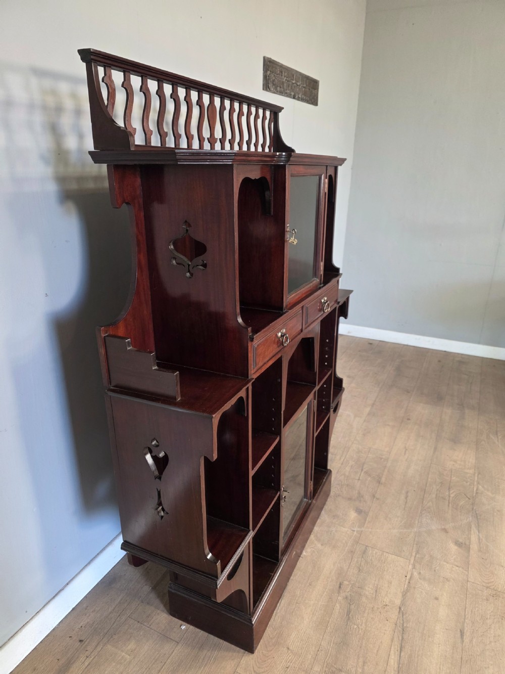 An antique dark mahogany Arts and Crafts display bookcase featuring a top galleried balustrade, open side shelves with pierced cut-outs, two glazed cabinet doors, and two middle drawers. View VII