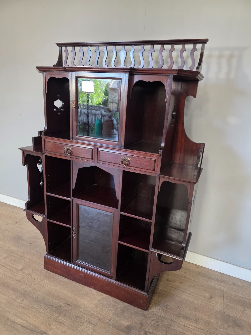 An antique dark mahogany Arts and Crafts display bookcase featuring a top galleried balustrade, open side shelves with pierced cut-outs, two glazed cabinet doors, and two middle drawers. View VIII