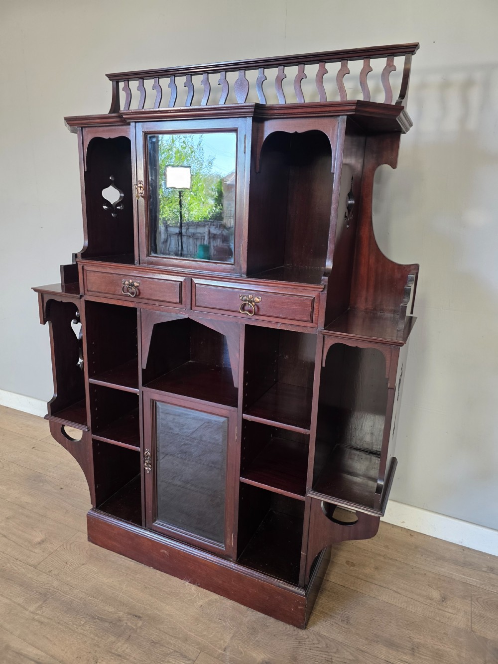 An antique dark mahogany Arts and Crafts display bookcase featuring a top galleried balustrade, open side shelves with pierced cut-outs, two glazed cabinet doors, and two middle drawers. View XI