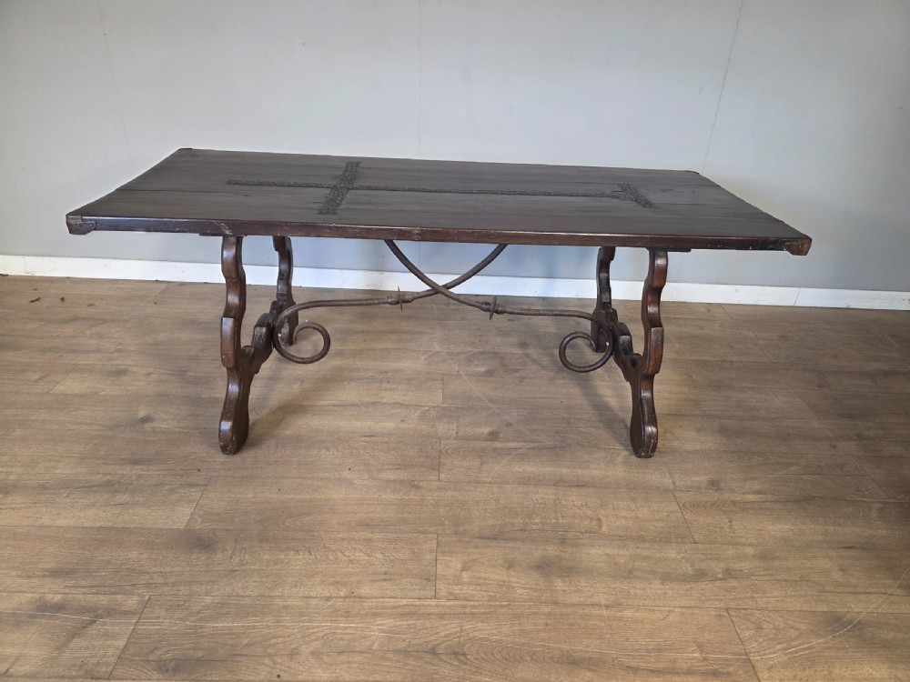 A dark brown oak refectory table featuring shaped trestle legs and a hand-forged wrought iron scrollwork stretcher beneath a rectangular top.