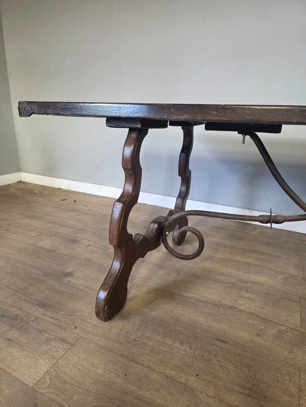 A dark brown oak refectory table featuring shaped trestle legs and a hand-forged wrought iron scrollwork stretcher beneath a rectangular top. View VII