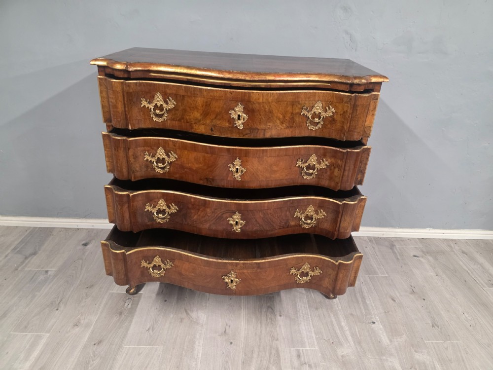 An 18th-century walnut chest of drawers featuring a serpentine front, four drawers, ornate brass handles, and short cabriole legs. View VI