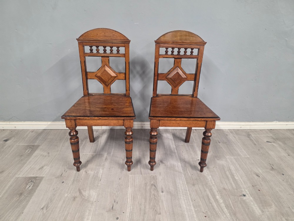 A pair of Victorian solid oak hall chairs featuring spindle pediments, central diamond back motifs, solid seats, and turned front legs. View II