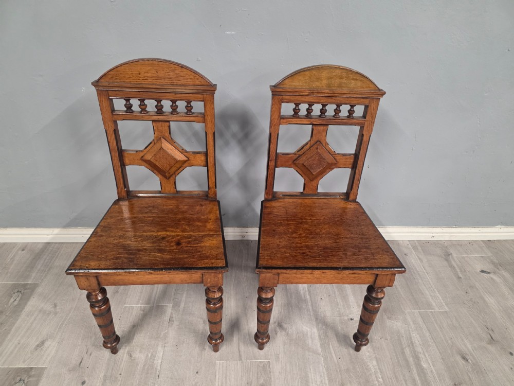 A pair of Victorian solid oak hall chairs featuring spindle pediments, central diamond back motifs, solid seats, and turned front legs. View III