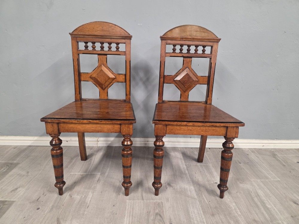 A pair of Victorian solid oak hall chairs featuring spindle pediments, central diamond back motifs, solid seats, and turned front legs. View IV