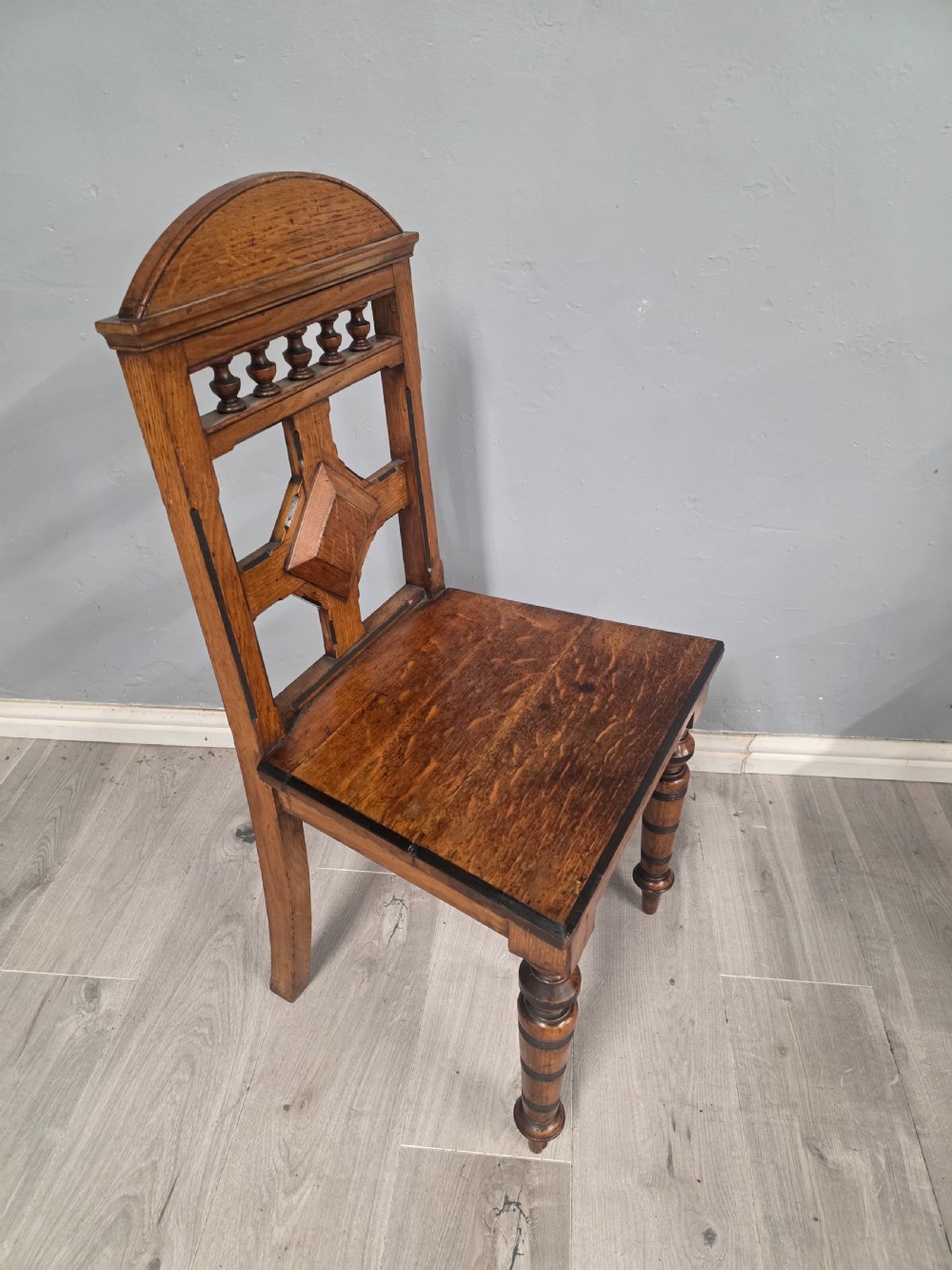 A pair of Victorian solid oak hall chairs featuring spindle pediments, central diamond back motifs, solid seats, and turned front legs. View VI