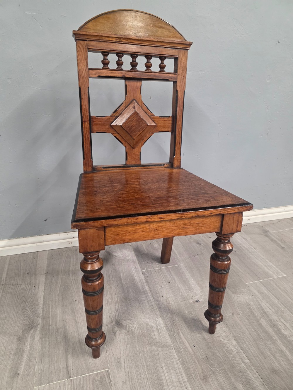 A pair of Victorian solid oak hall chairs featuring spindle pediments, central diamond back motifs, solid seats, and turned front legs. View X