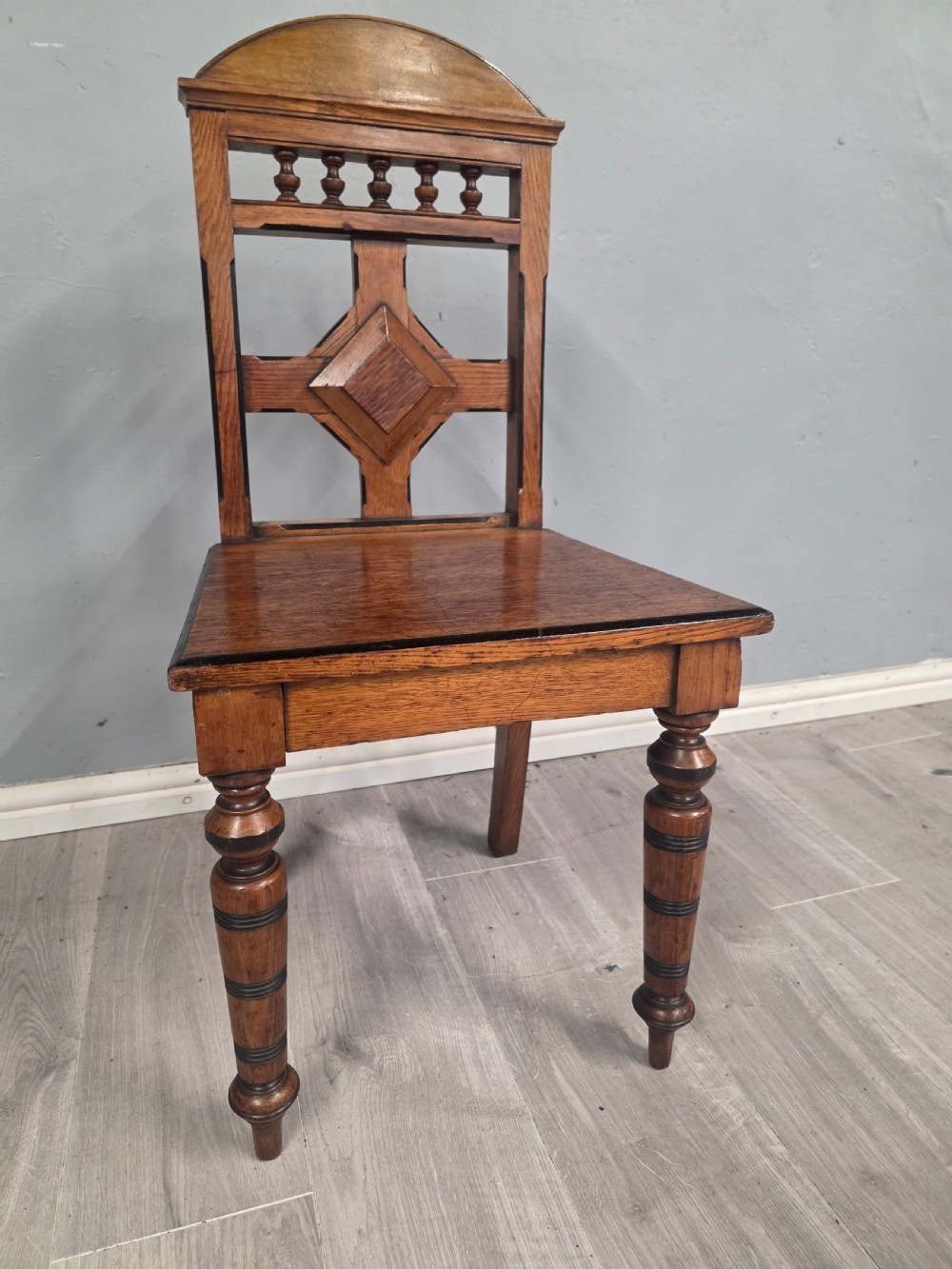 A pair of Victorian solid oak hall chairs featuring spindle pediments, central diamond back motifs, solid seats, and turned front legs. View XI