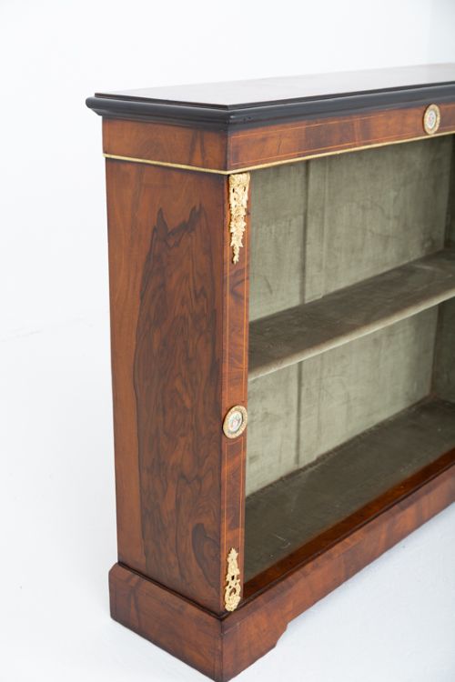 A Victorian burr walnut open bookcase featuring two open shelves, ebonised top trim, gilt brass mounts, and ceramic plaque details. View VI