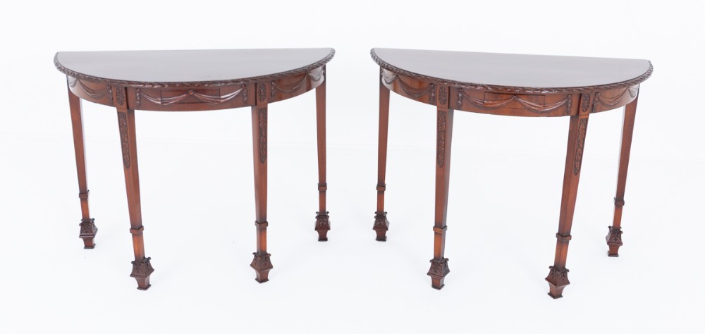 A pair of demilune mahogany console tables with carved drapery aprons and slender tapered legs ending in decorative feet.