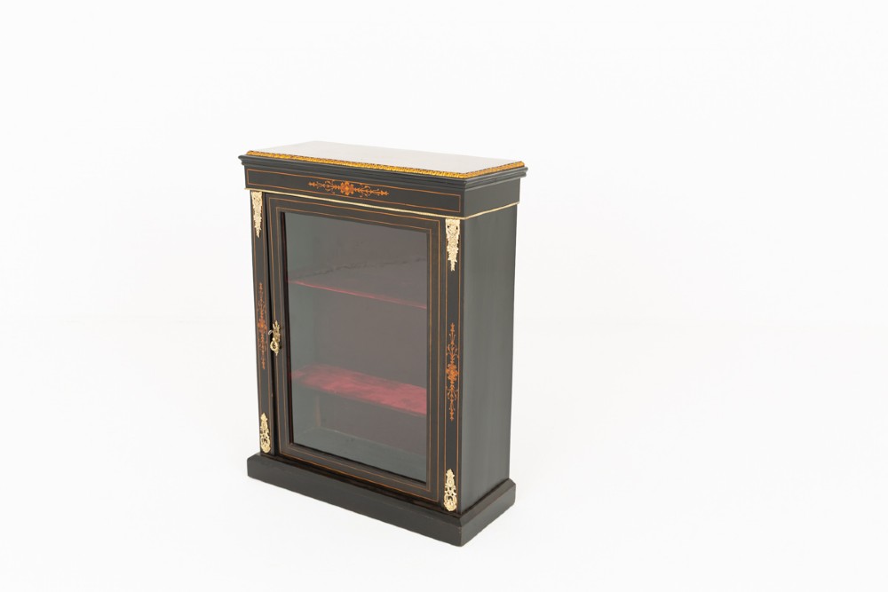 An ebonised Victorian pier cabinet with gilt brass mounts, detailed wood inlay, a single glazed door, and red-lined interior shelves. View IV