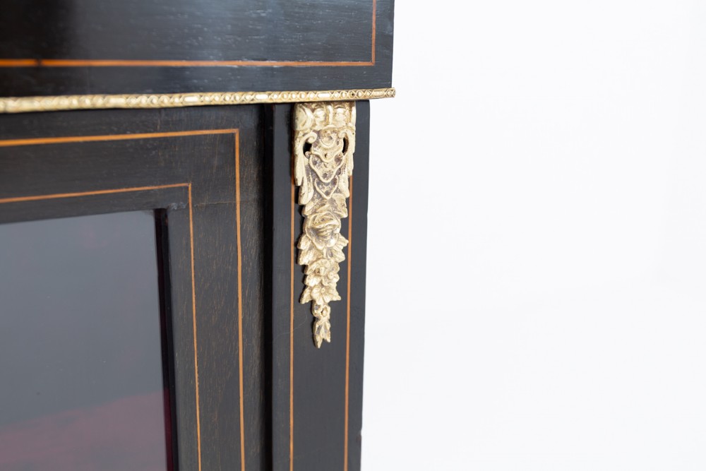 An ebonised Victorian pier cabinet with gilt brass mounts, detailed wood inlay, a single glazed door, and red-lined interior shelves. View VI