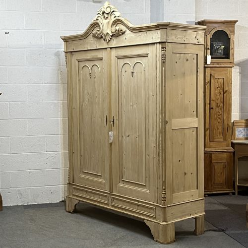 fig 1. A large late nineteenth-century natural pine double wardrobe featuring a carved scroll pediment, arched door mouldings, and a flat plinth base.