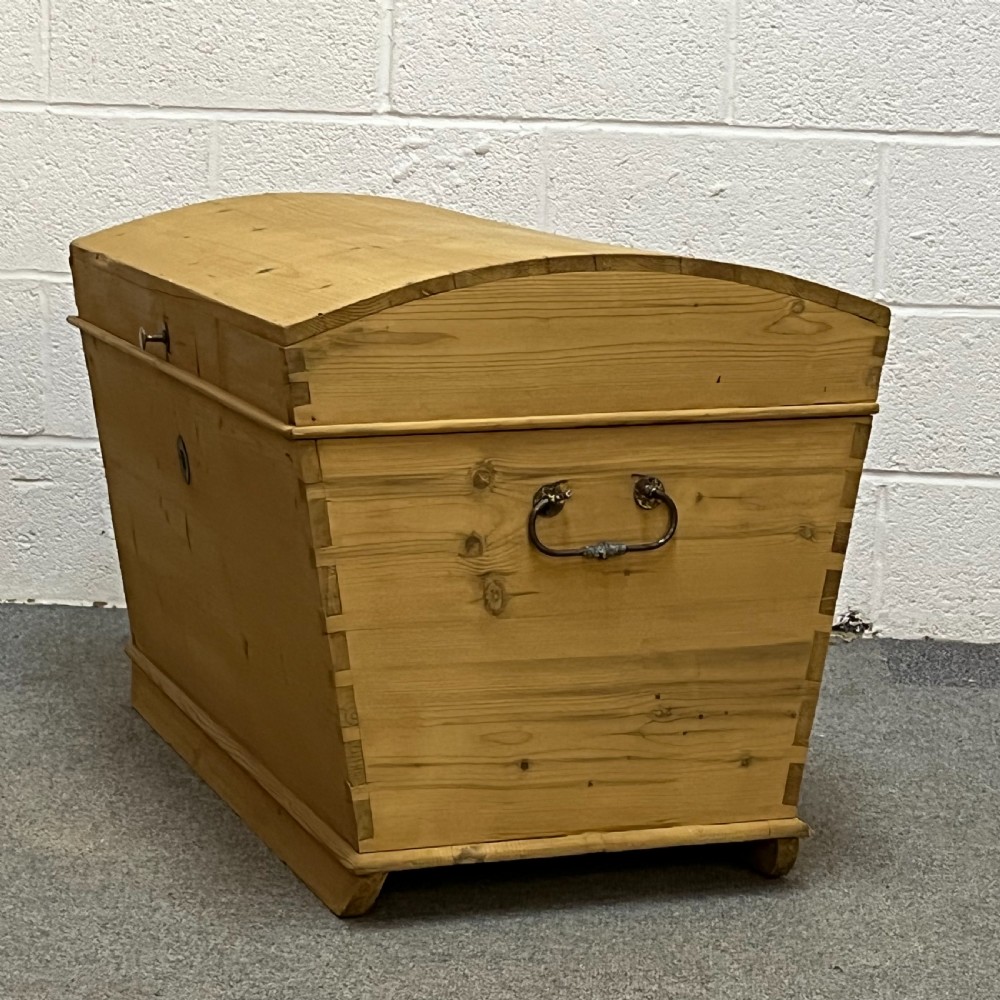 A solid honey-toned pine dome top trunk with visible corner dovetail joints, metal side carrying handles, and a central keyhole escutcheon. View II