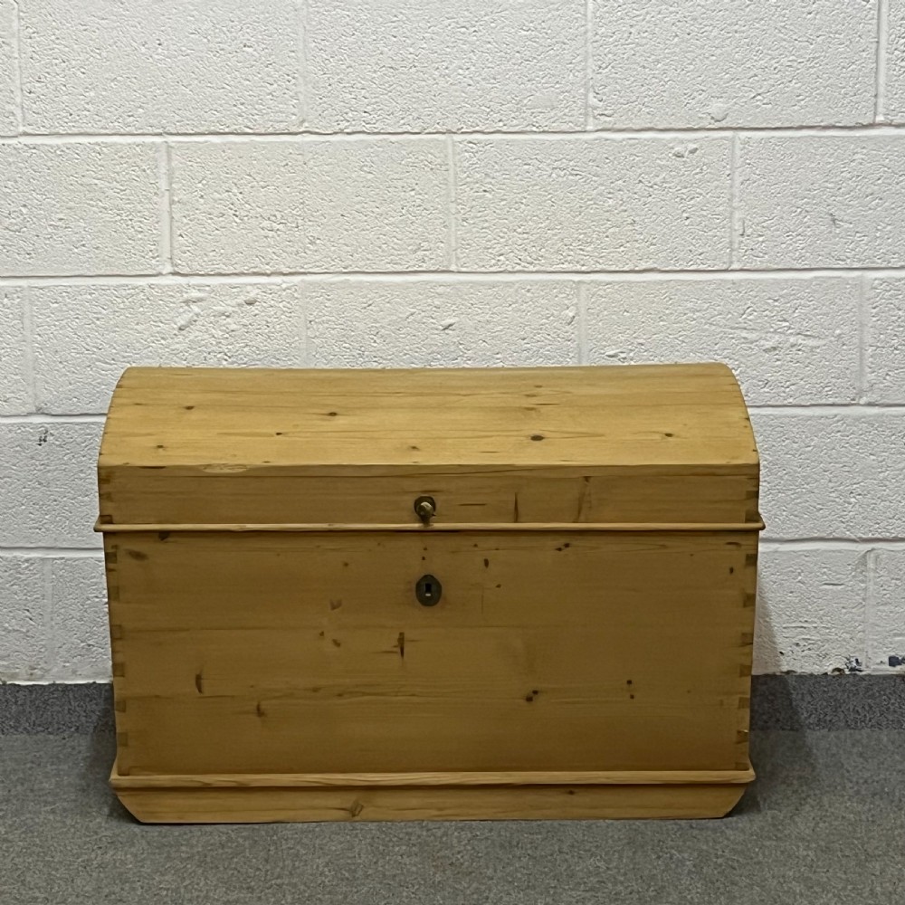 A solid honey-toned pine dome top trunk with visible corner dovetail joints, metal side carrying handles, and a central keyhole escutcheon. View V