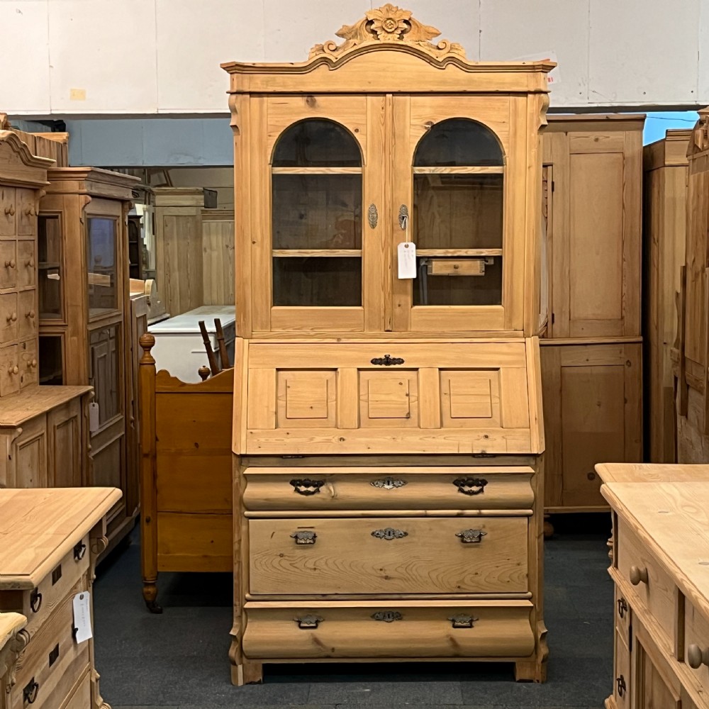 A tall antique pine secretaire bookcase featuring an upper glazed cabinet with arched doors, a fall-front writing surface, and a lower chest of drawers with decorative handles.