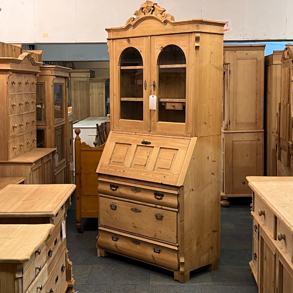 A tall antique pine secretaire bookcase featuring an upper glazed cabinet with arched doors, a fall-front writing surface, and a lower chest of drawers with decorative handles. View III