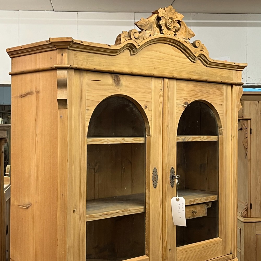A tall antique pine secretaire bookcase featuring an upper glazed cabinet with arched doors, a fall-front writing surface, and a lower chest of drawers with decorative handles. View VI