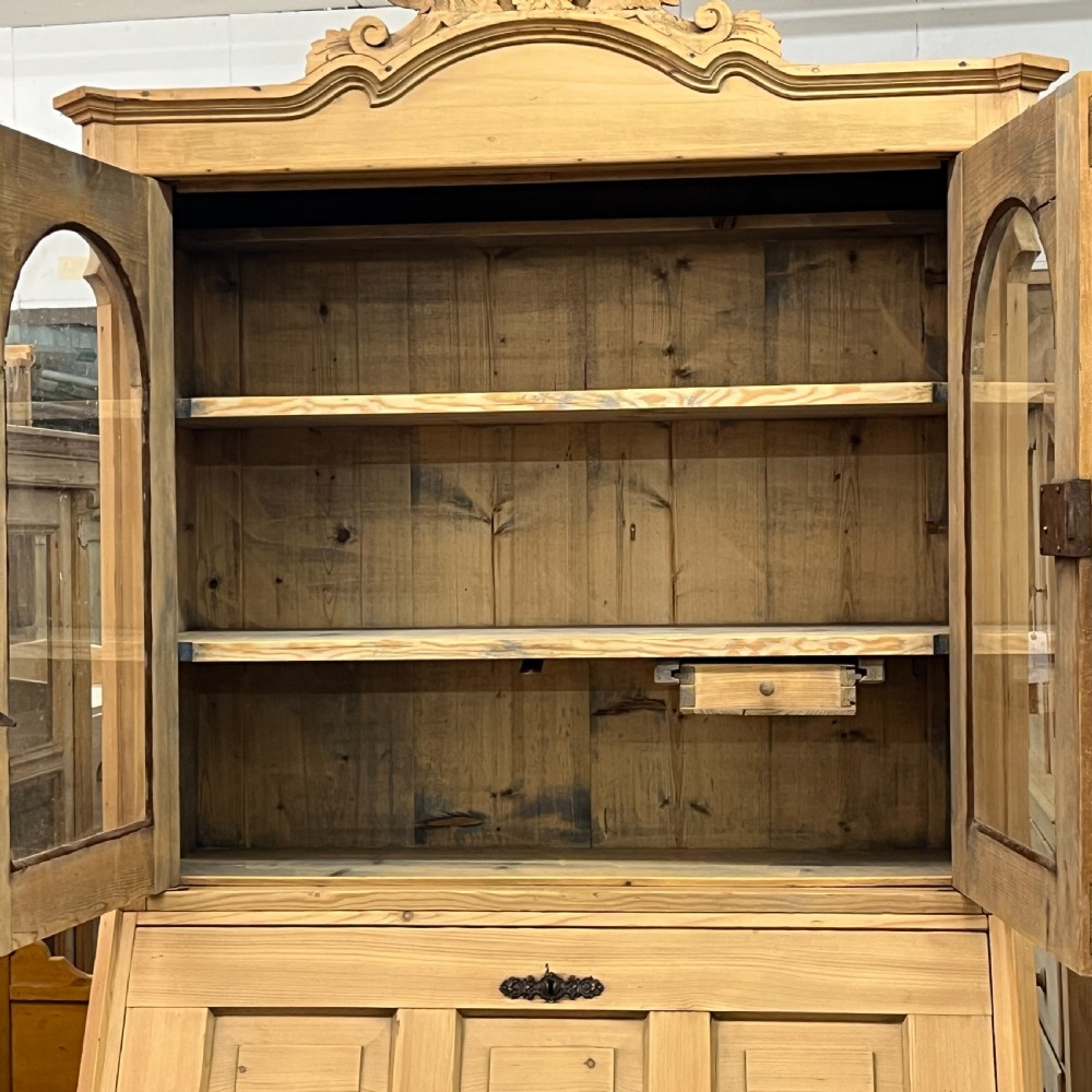 A tall antique pine secretaire bookcase featuring an upper glazed cabinet with arched doors, a fall-front writing surface, and a lower chest of drawers with decorative handles. View X