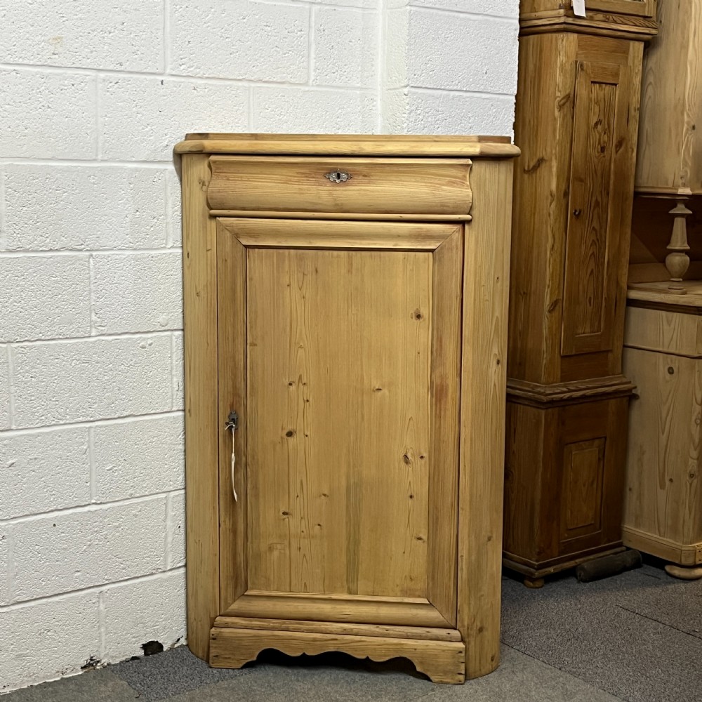 A natural stripped pine freestanding corner cupboard featuring a top drawer, a single panel door with keyhole escutcheon, and a shaped apron base.