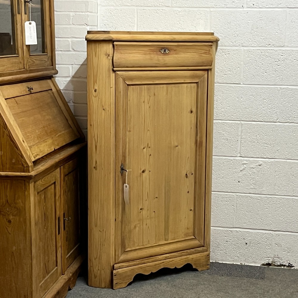 A natural stripped pine freestanding corner cupboard featuring a top drawer, a single panel door with keyhole escutcheon, and a shaped apron base. View II