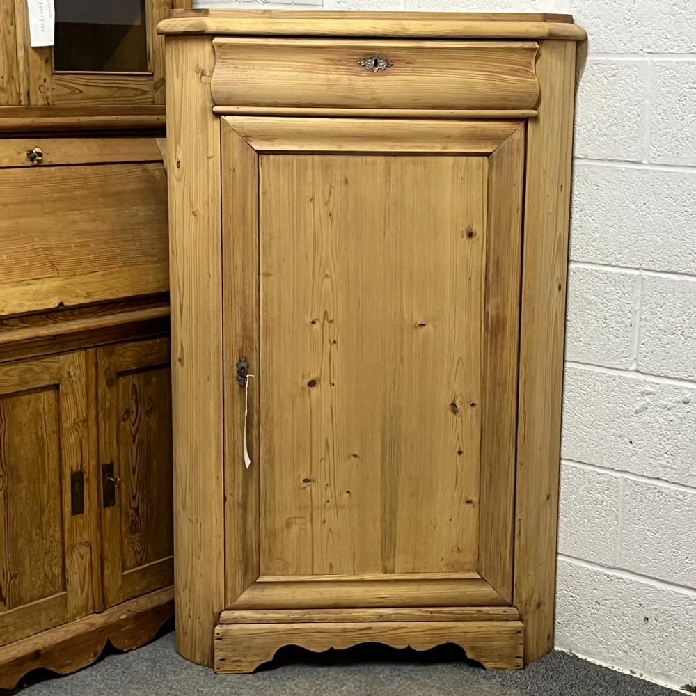 A natural stripped pine freestanding corner cupboard featuring a top drawer, a single panel door with keyhole escutcheon, and a shaped apron base. View IV