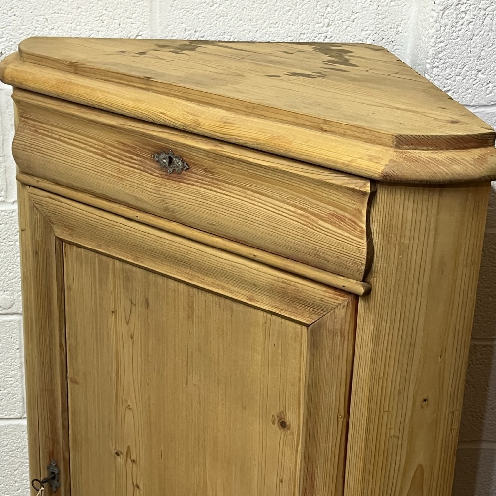 A natural stripped pine freestanding corner cupboard featuring a top drawer, a single panel door with keyhole escutcheon, and a shaped apron base. View V