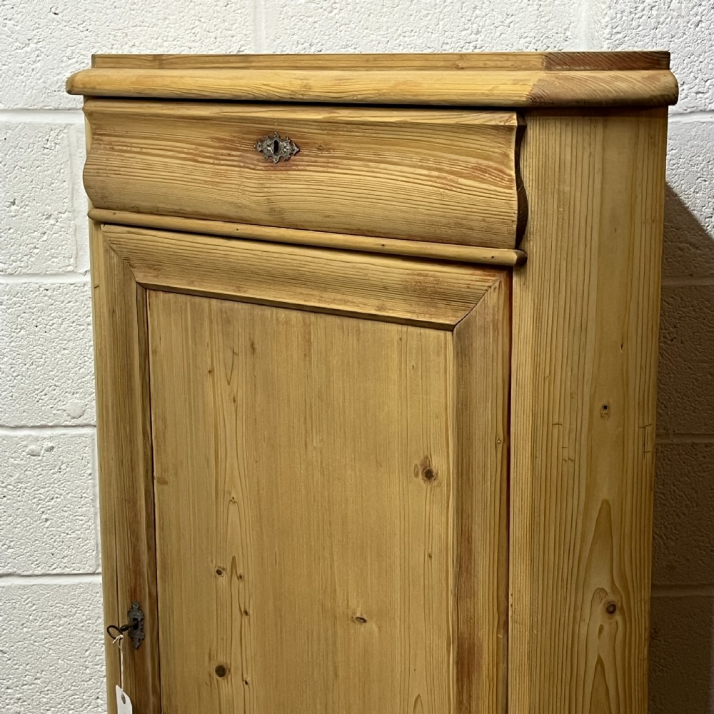 A natural stripped pine freestanding corner cupboard featuring a top drawer, a single panel door with keyhole escutcheon, and a shaped apron base. View VI