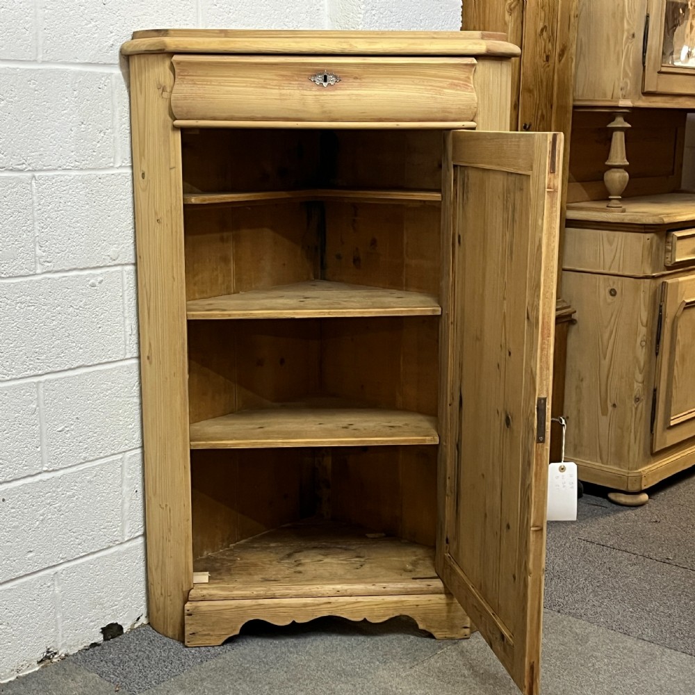 A natural stripped pine freestanding corner cupboard featuring a top drawer, a single panel door with keyhole escutcheon, and a shaped apron base. View VIII