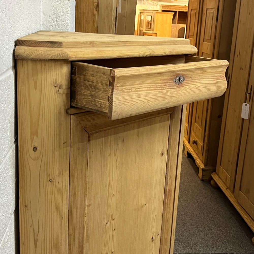 A natural stripped pine freestanding corner cupboard featuring a top drawer, a single panel door with keyhole escutcheon, and a shaped apron base. View X