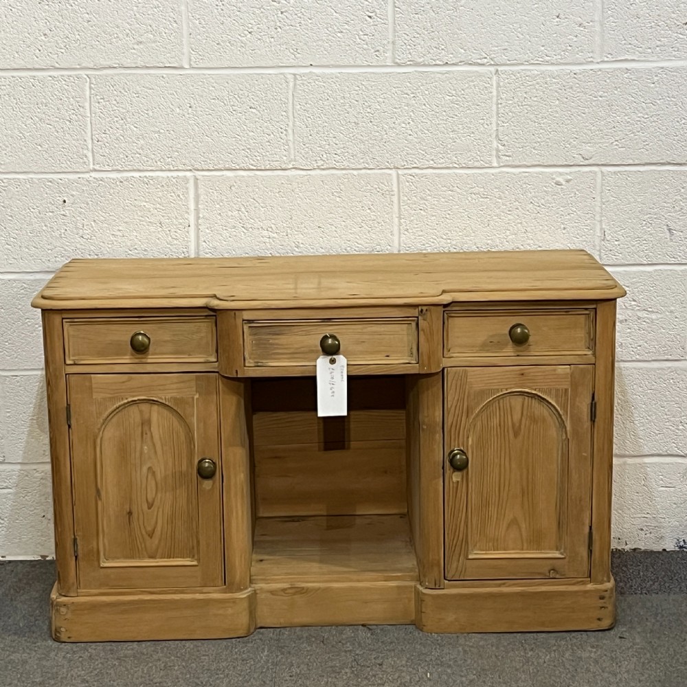 A Victorian pine kneehole writing desk featuring three upper drawers, two arched cupboard doors, metal knobs, and a solid plinth base.