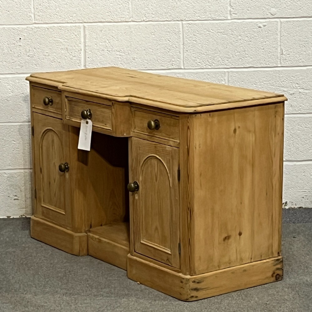 A Victorian pine kneehole writing desk featuring three upper drawers, two arched cupboard doors, metal knobs, and a solid plinth base. View II