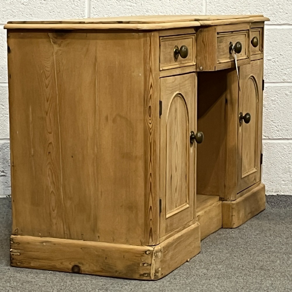 A Victorian pine kneehole writing desk featuring three upper drawers, two arched cupboard doors, metal knobs, and a solid plinth base. View III