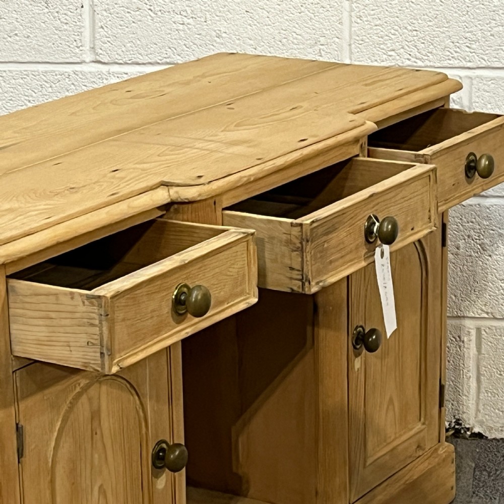 A Victorian pine kneehole writing desk featuring three upper drawers, two arched cupboard doors, metal knobs, and a solid plinth base. View V