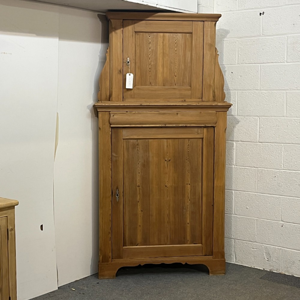 A tall two-tier antique pine corner cupboard with single panelled upper and lower doors and side scroll details.