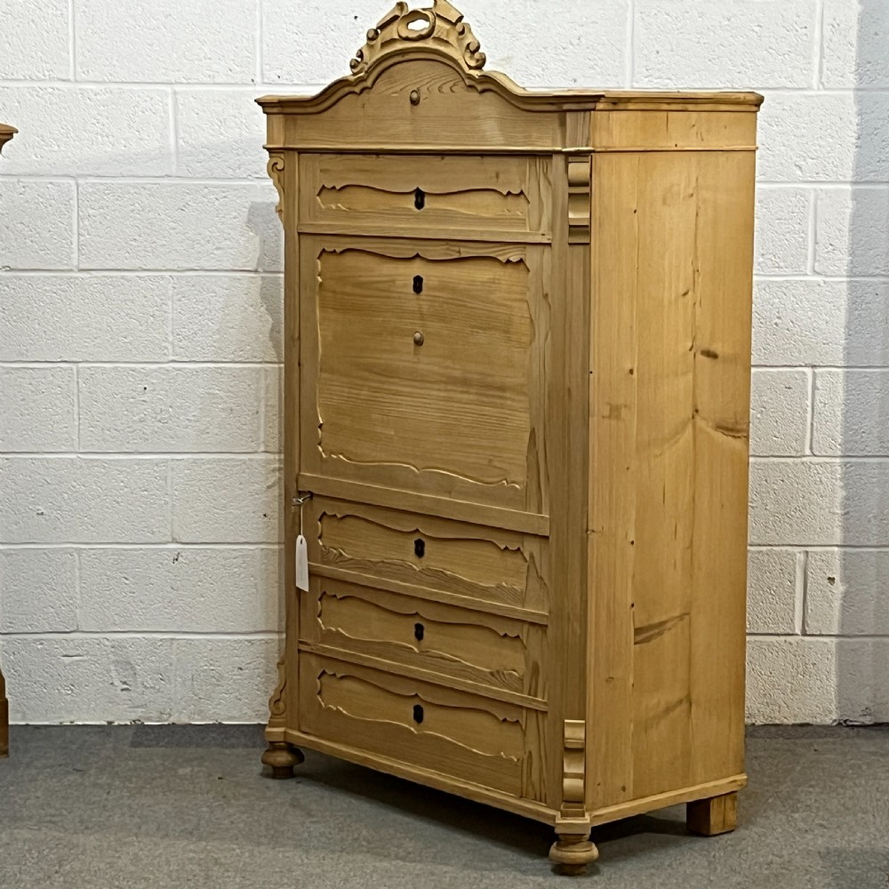 A tall antique pine single-door cupboard featuring a decorative carved cresting, moulding styled like drawers, central keyholes, and turned feet. View II