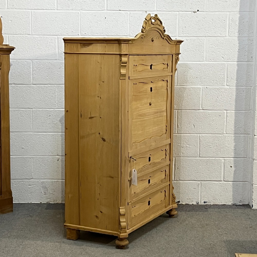 A tall antique pine single-door cupboard featuring a decorative carved cresting, moulding styled like drawers, central keyholes, and turned feet. View III