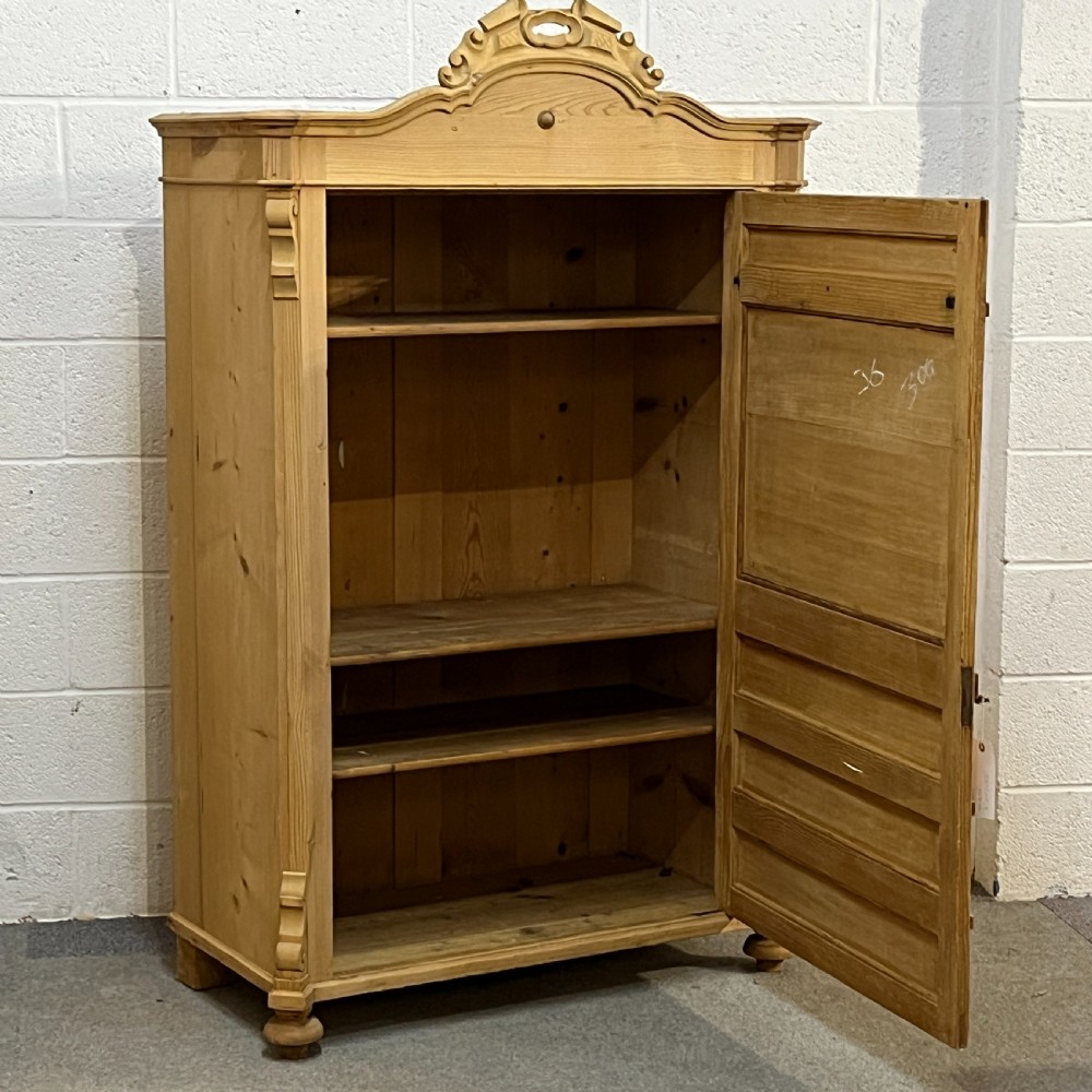 A tall antique pine single-door cupboard featuring a decorative carved cresting, moulding styled like drawers, central keyholes, and turned feet. View V
