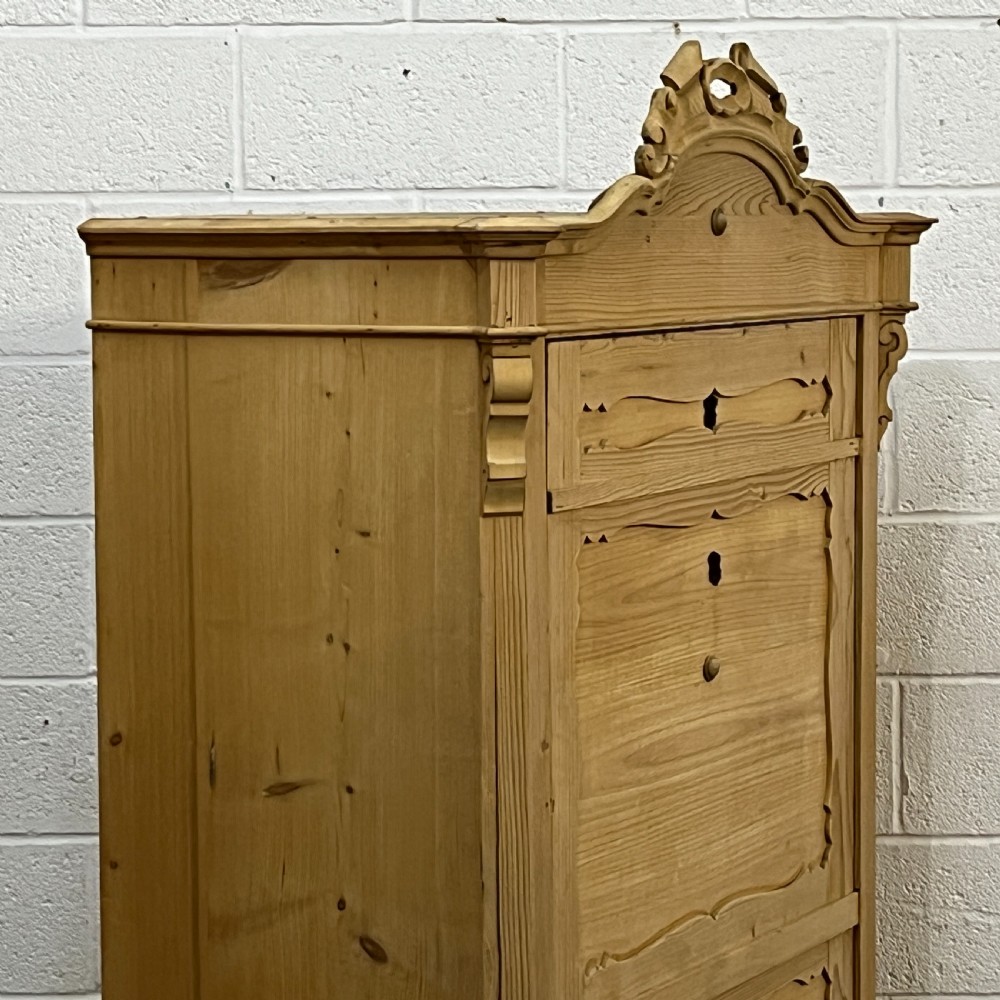 A tall antique pine single-door cupboard featuring a decorative carved cresting, moulding styled like drawers, central keyholes, and turned feet. View VI