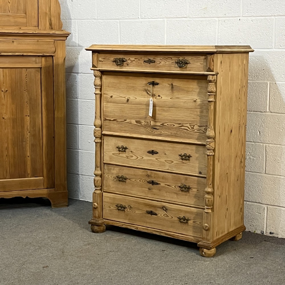 An antique pine secretaire desk featuring a fall-front writing compartment above multiple drawers, decorated with turned side columns and dark metal handles.