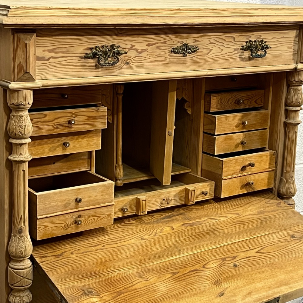 An antique pine secretaire desk featuring a fall-front writing compartment above multiple drawers, decorated with turned side columns and dark metal handles. View V
