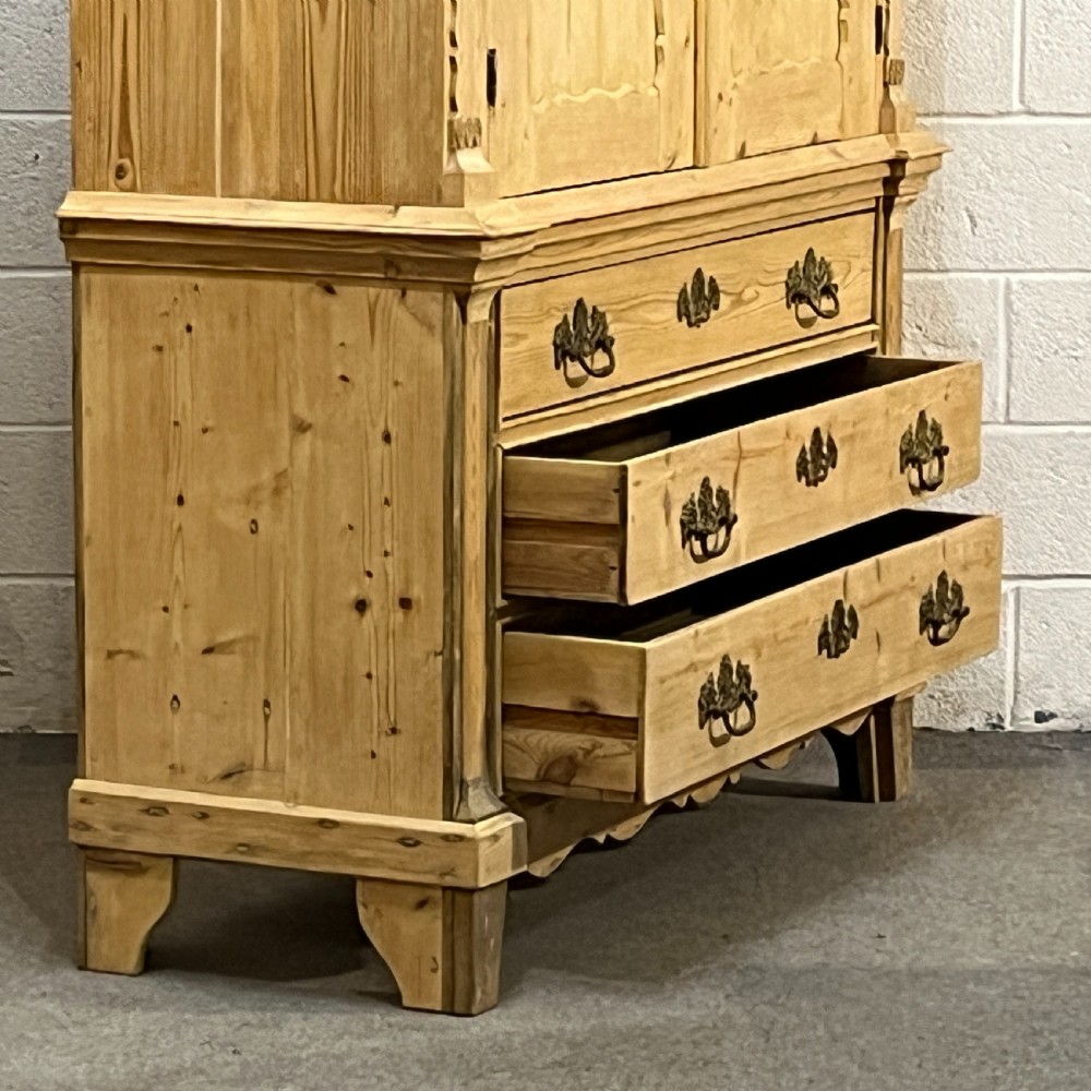 A tall two-part natural pine linen press featuring a double-door top cupboard over three drawers with brass drop handles. View V