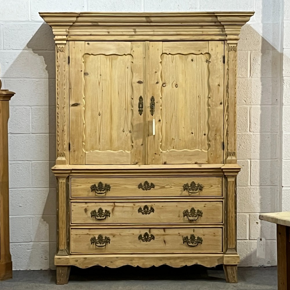 A tall two-part natural pine linen press featuring a double-door top cupboard over three drawers with brass drop handles. View VI