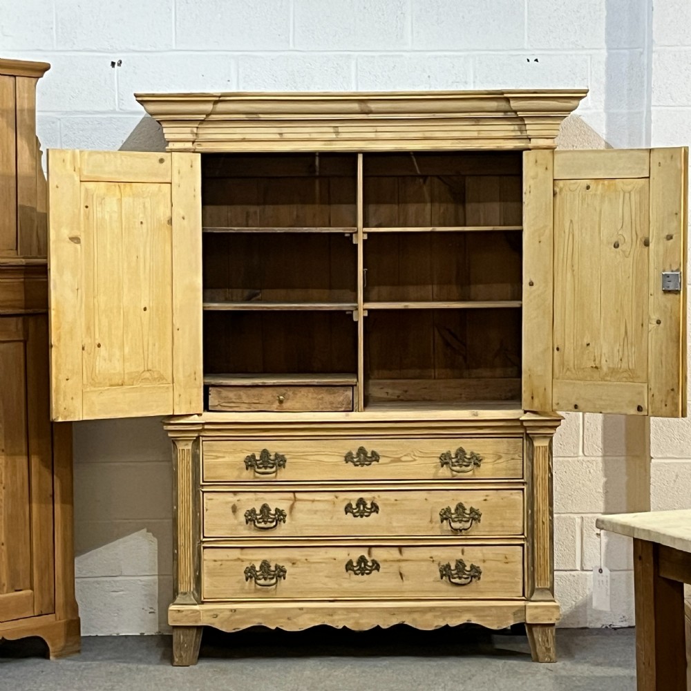 A tall two-part natural pine linen press featuring a double-door top cupboard over three drawers with brass drop handles. View VII